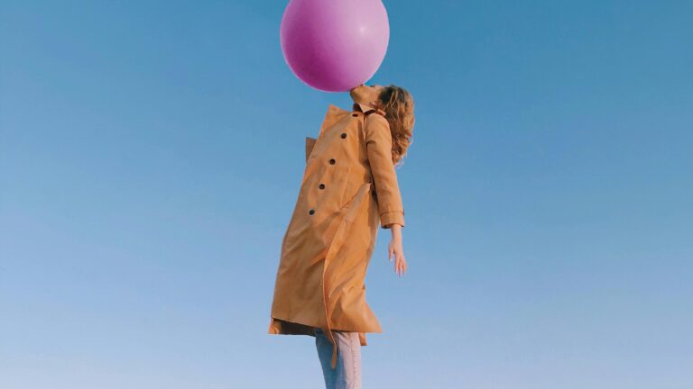Side view of woman wearing trendy clothes with trench coat flying in sky and blowing enormous balloon